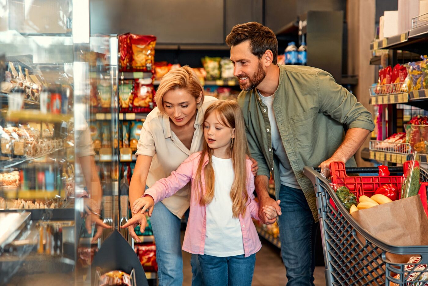 Junge, glückliche Familie mit einem Kind kauft Lebensmittel in einem Geschäft, sie stehen vor Regalen mit ihrem Einkaufswagen und sind fröhlich und harmonisch