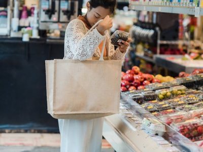 Junge Frau mit Stofftasche prüft ein Produkt in der Obst- und Gemüseabteilung eines modernen Supermarkts